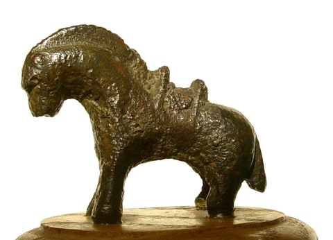 Bronzen paardje met de kop naar links, kamvormige manen en een aflopende rug, op een houten ondergrond.