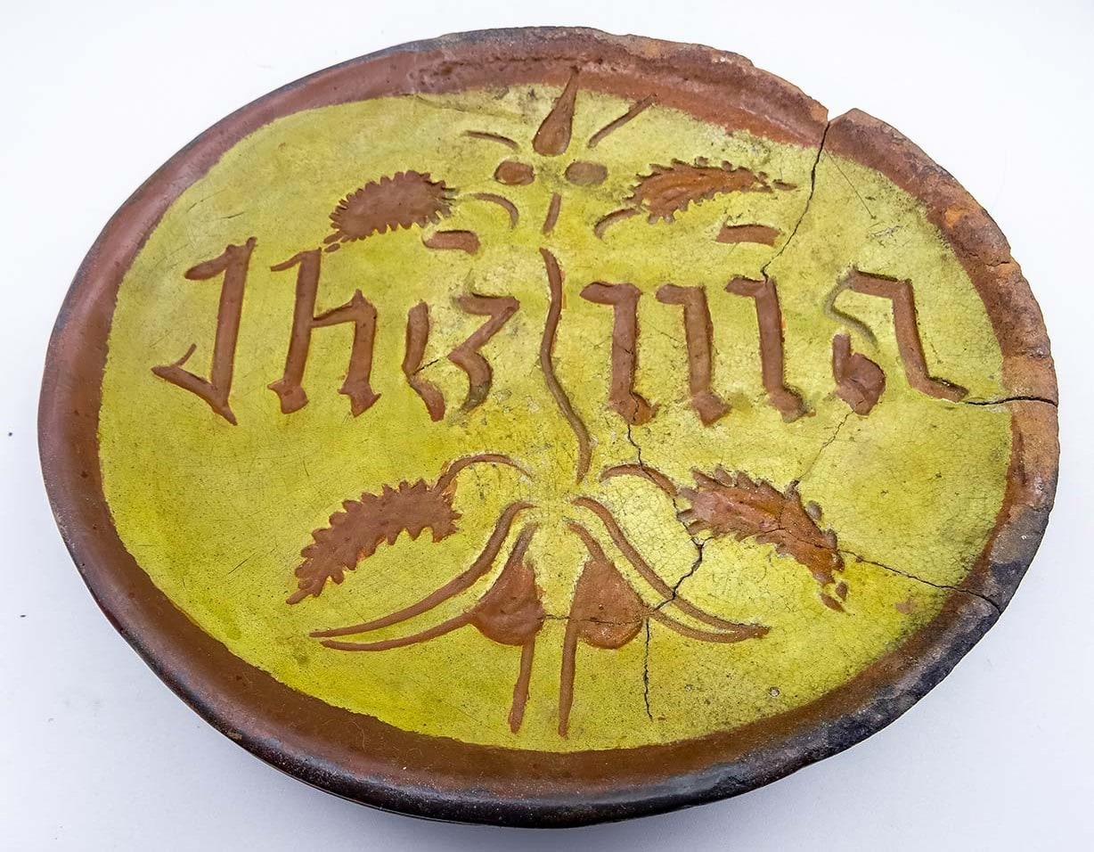 Roodbakkende voetschaal met gele binnenkant en de ingekraste letters IHS en MA rond een lelie.